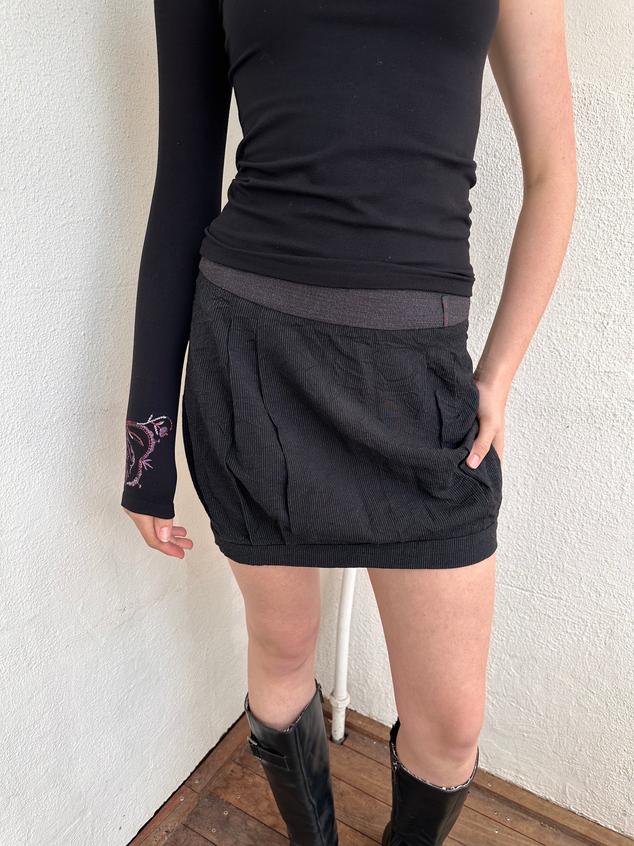 Person wearing a black outfit with a cop copine mini skirt, one shoulder top and boots standing on a light-coloured floor.