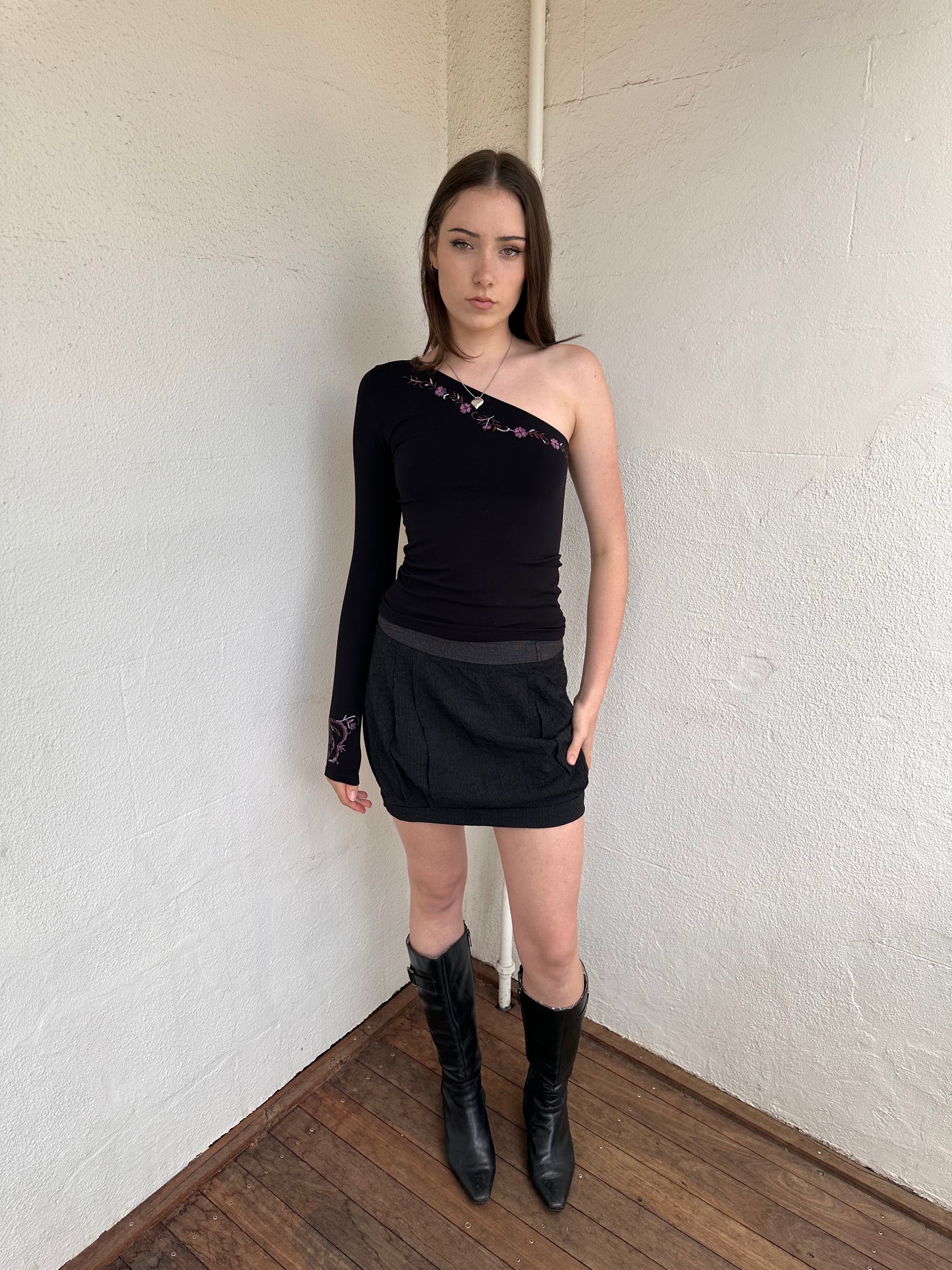 Person wearing a black outfit with a cop copine mini skirt, one shoulder top and boots standing on a light-coloured floor.