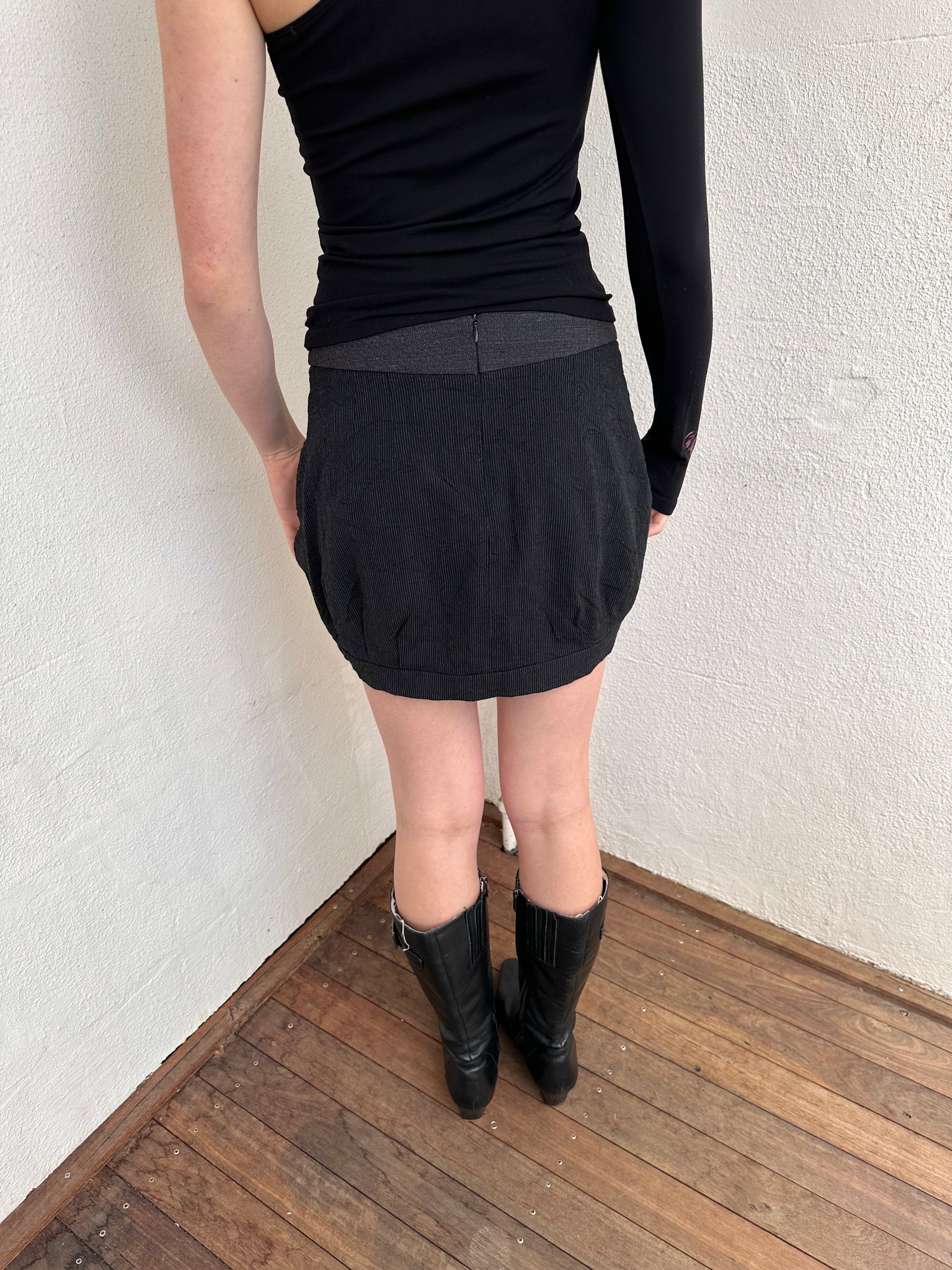 Person wearing a black outfit with a cop copine mini skirt, one shoulder top and boots standing on a light-coloured floor.