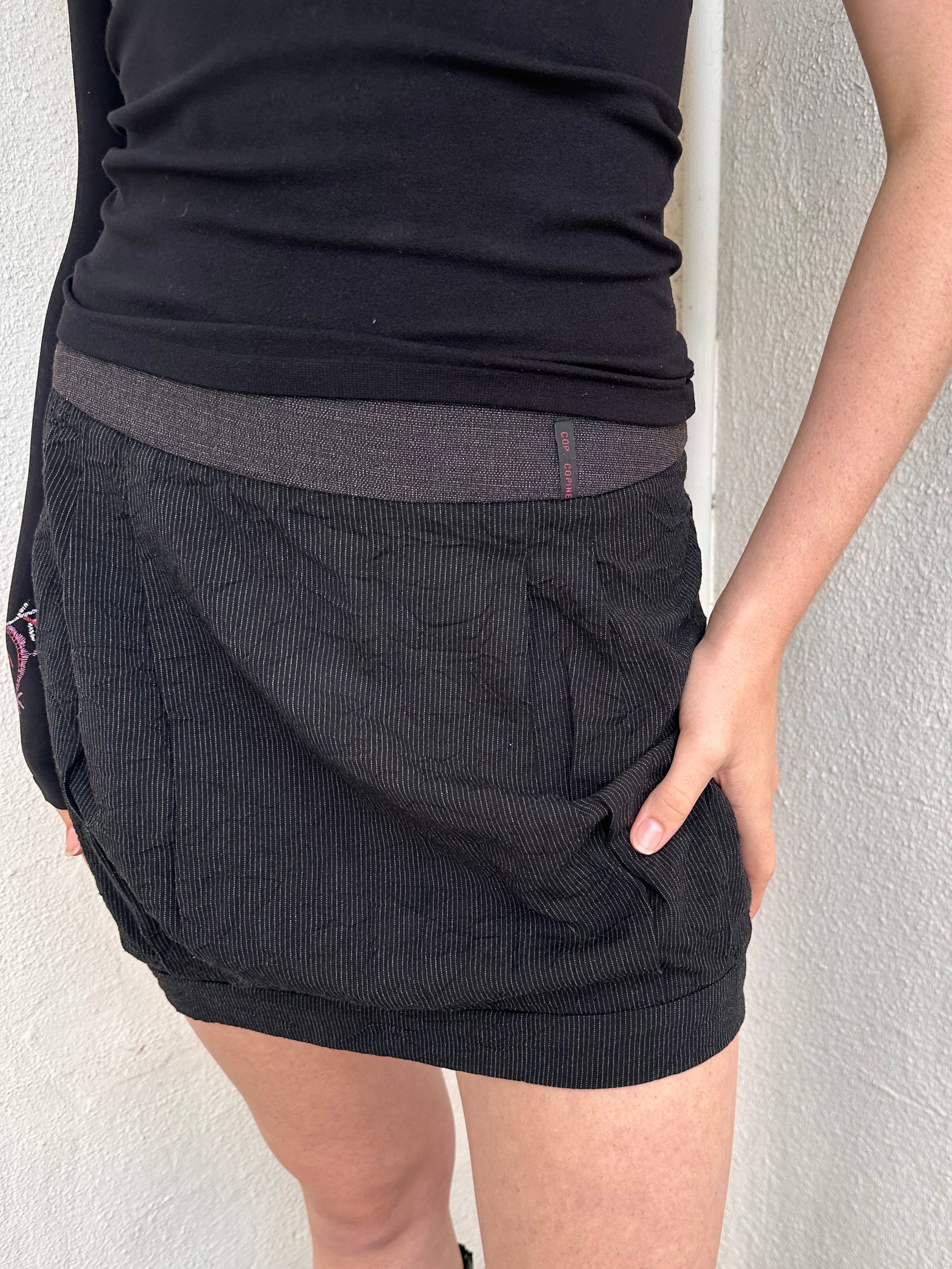 Person wearing a black outfit with a cop copine mini skirt, one shoulder top and boots standing on a light-coloured floor.