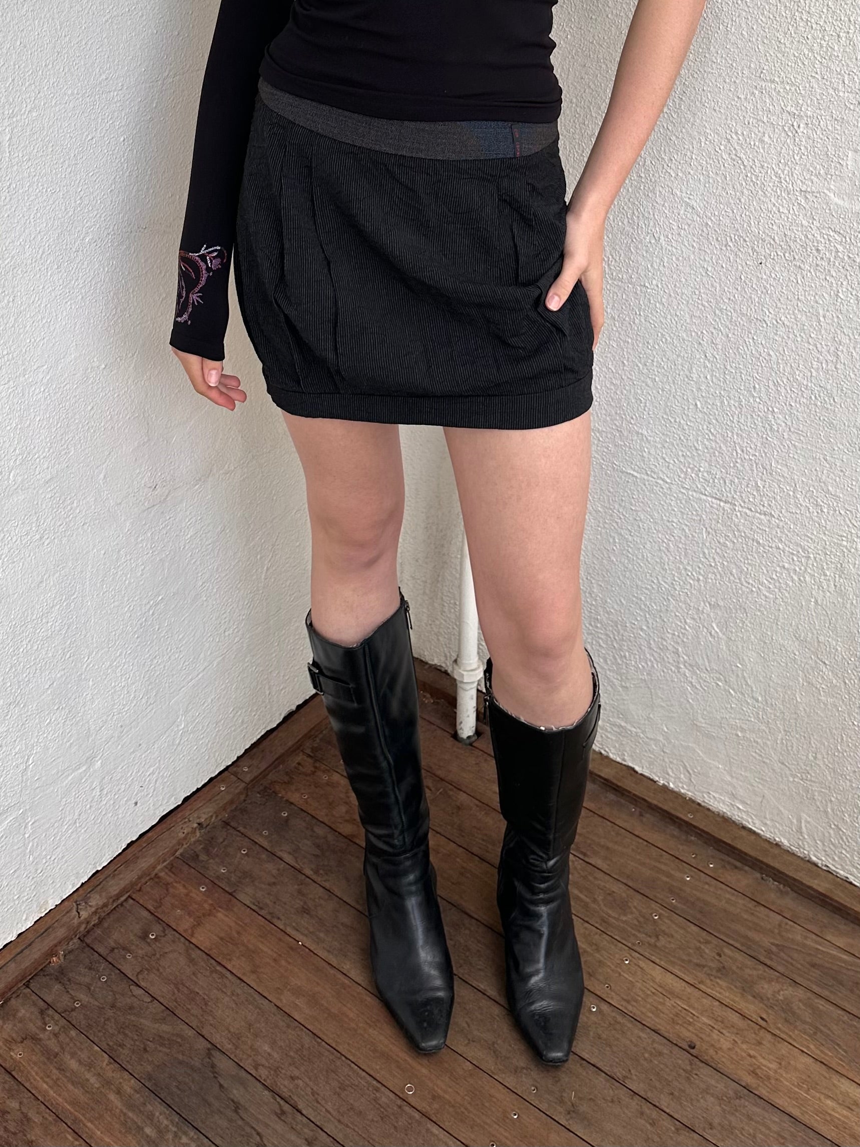 Person wearing a black outfit with a cop copine mini skirt, one shoulder top and boots standing on a light-coloured floor.