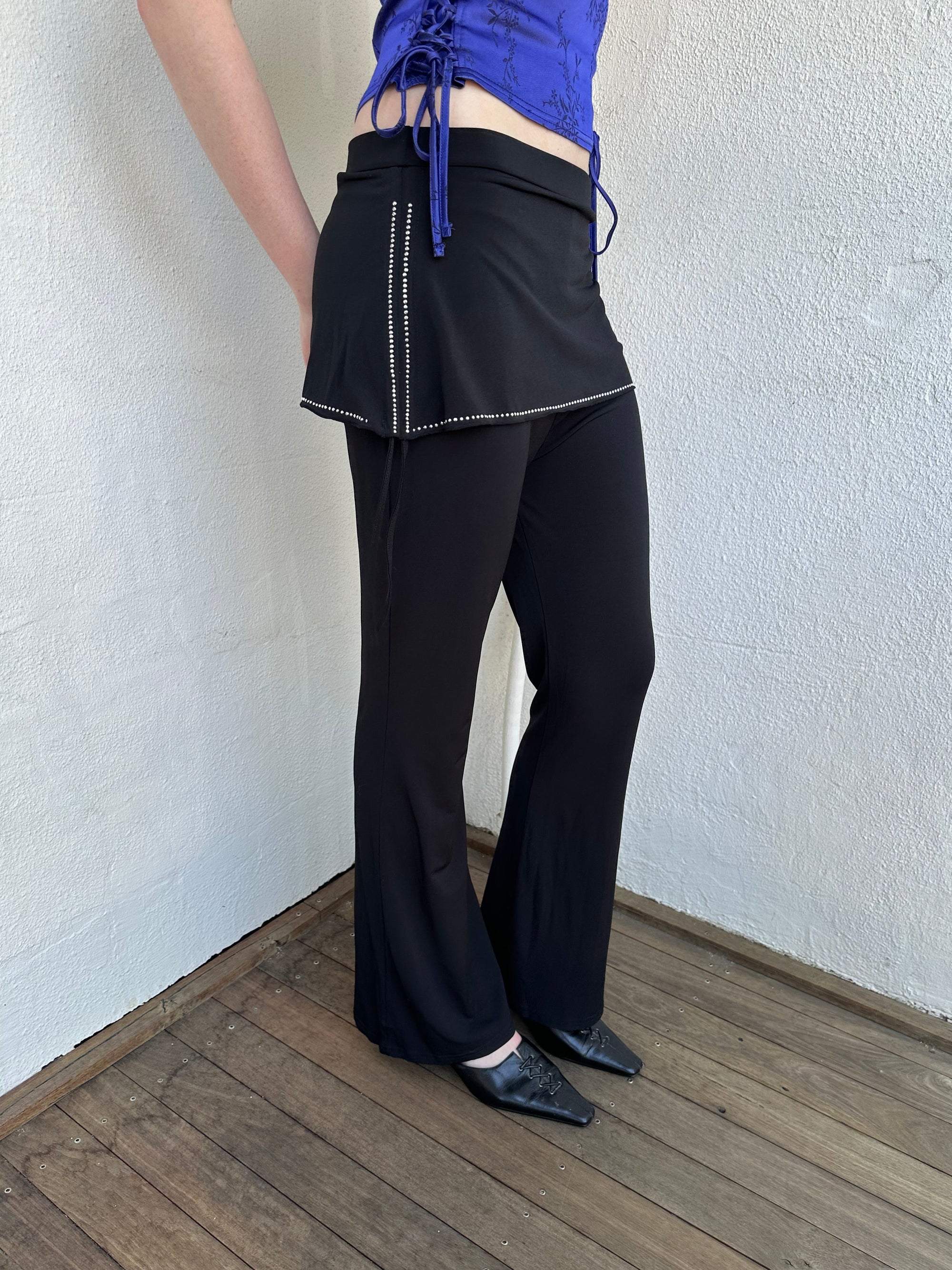 Woman Dressed in Y2K Black rhinestone detail skirt pants featuring a mini skirt attached over flared pants. 