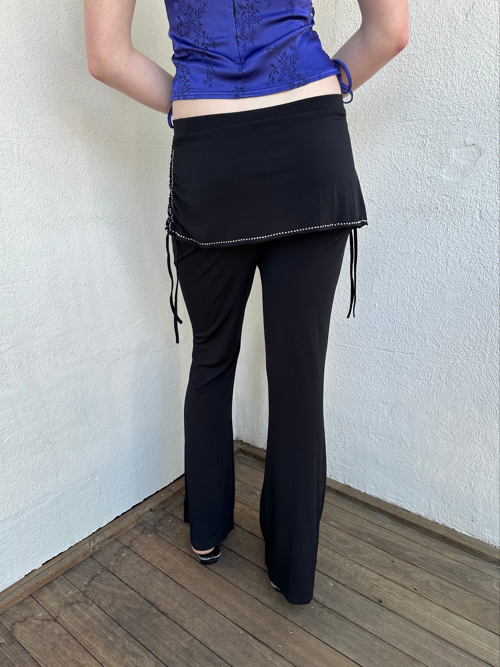Woman Dressed in Y2K Black rhinestone detail skirt pants featuring a mini skirt attached over flared pants. 