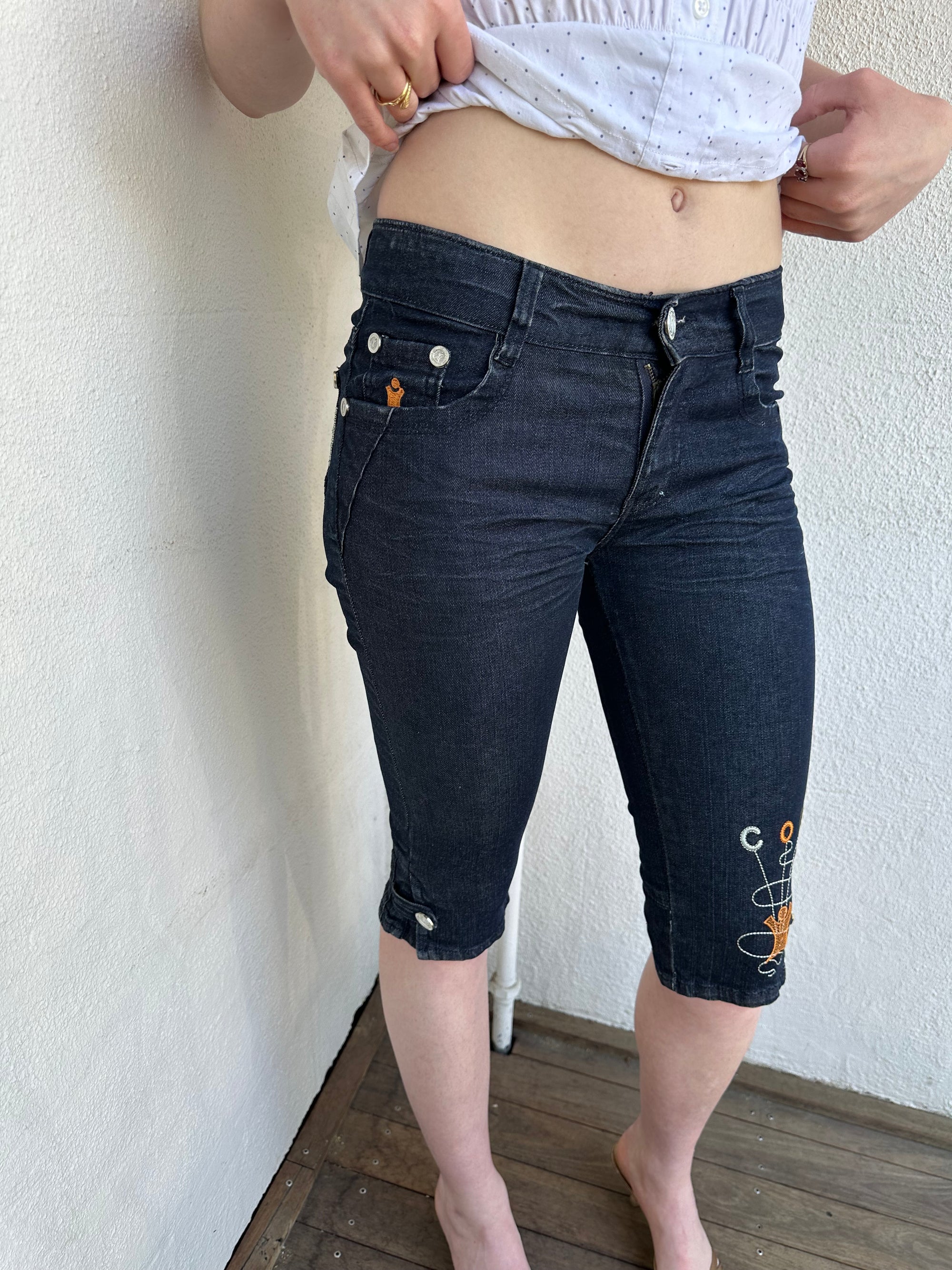 Woman Dressed in Y2K Cop Copine embroidered denim capris featuring dark denim with iconic orange and white Cop Copine logo embroidery.