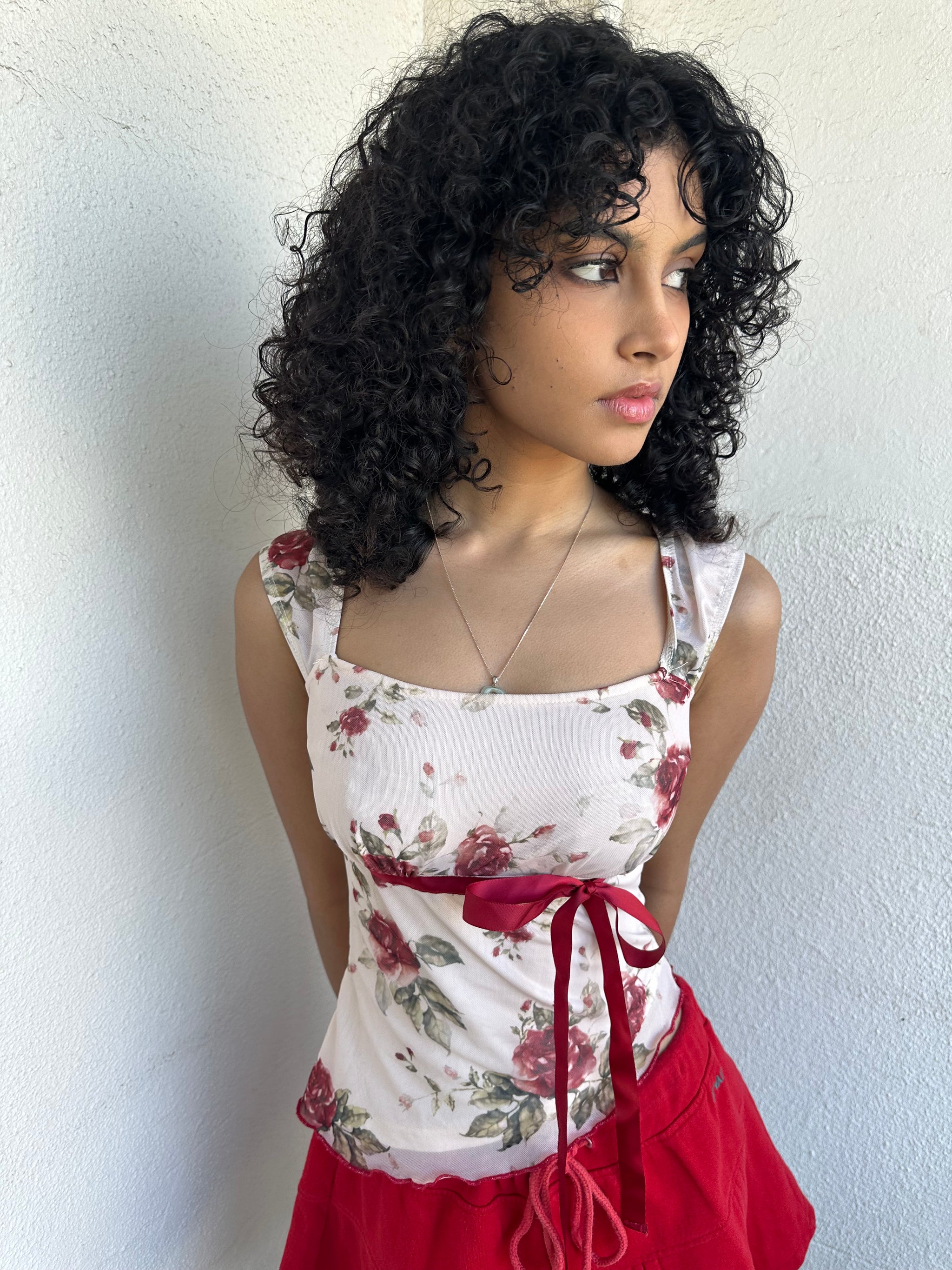 Woman wearing a Y2K 00s cream and red floral mesh tank, with an asymmetric hem and a red underbust ribbon finished with a bow. 
