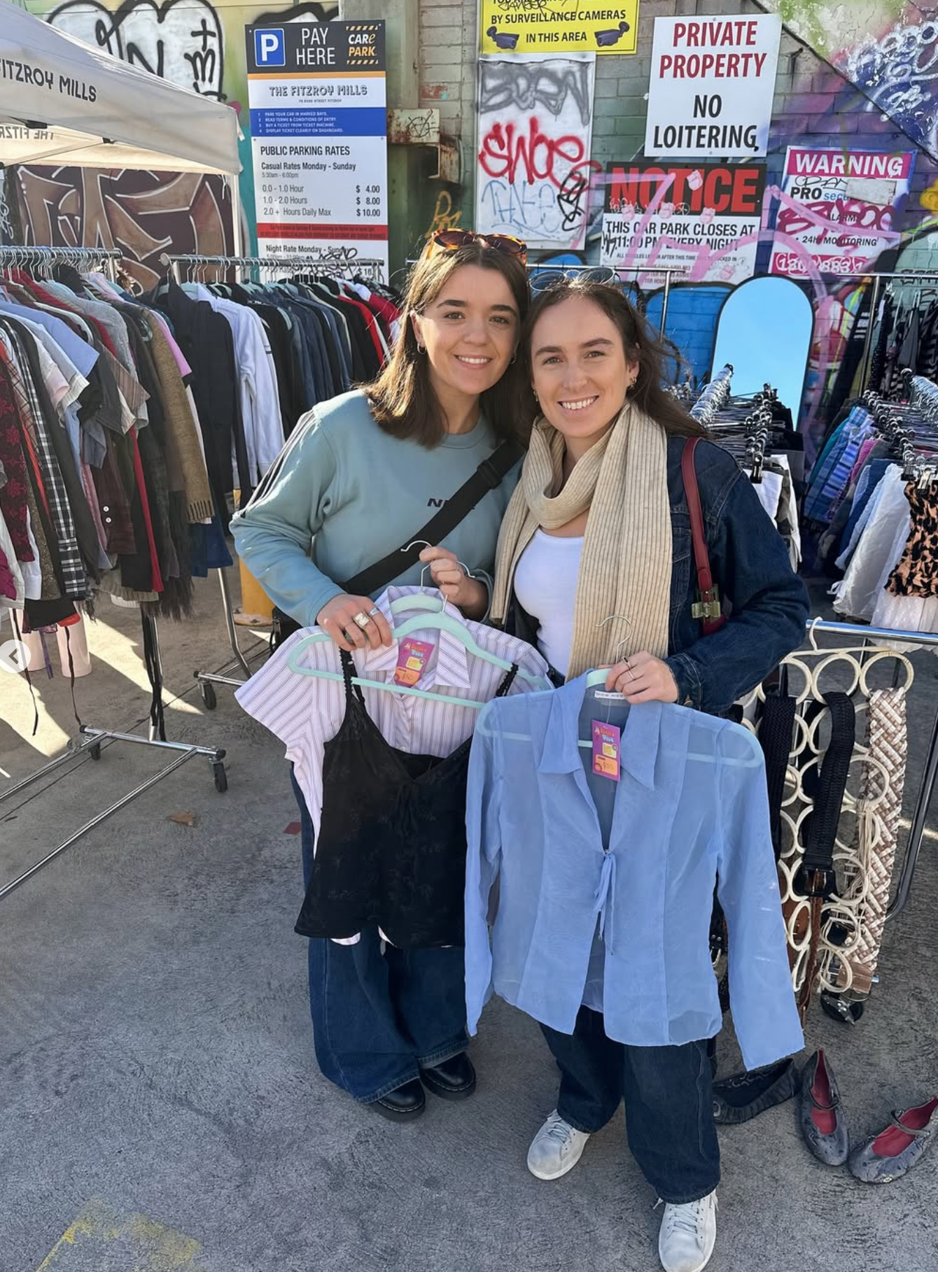 Two women at market holding clothing items with various signs and booths in the background.