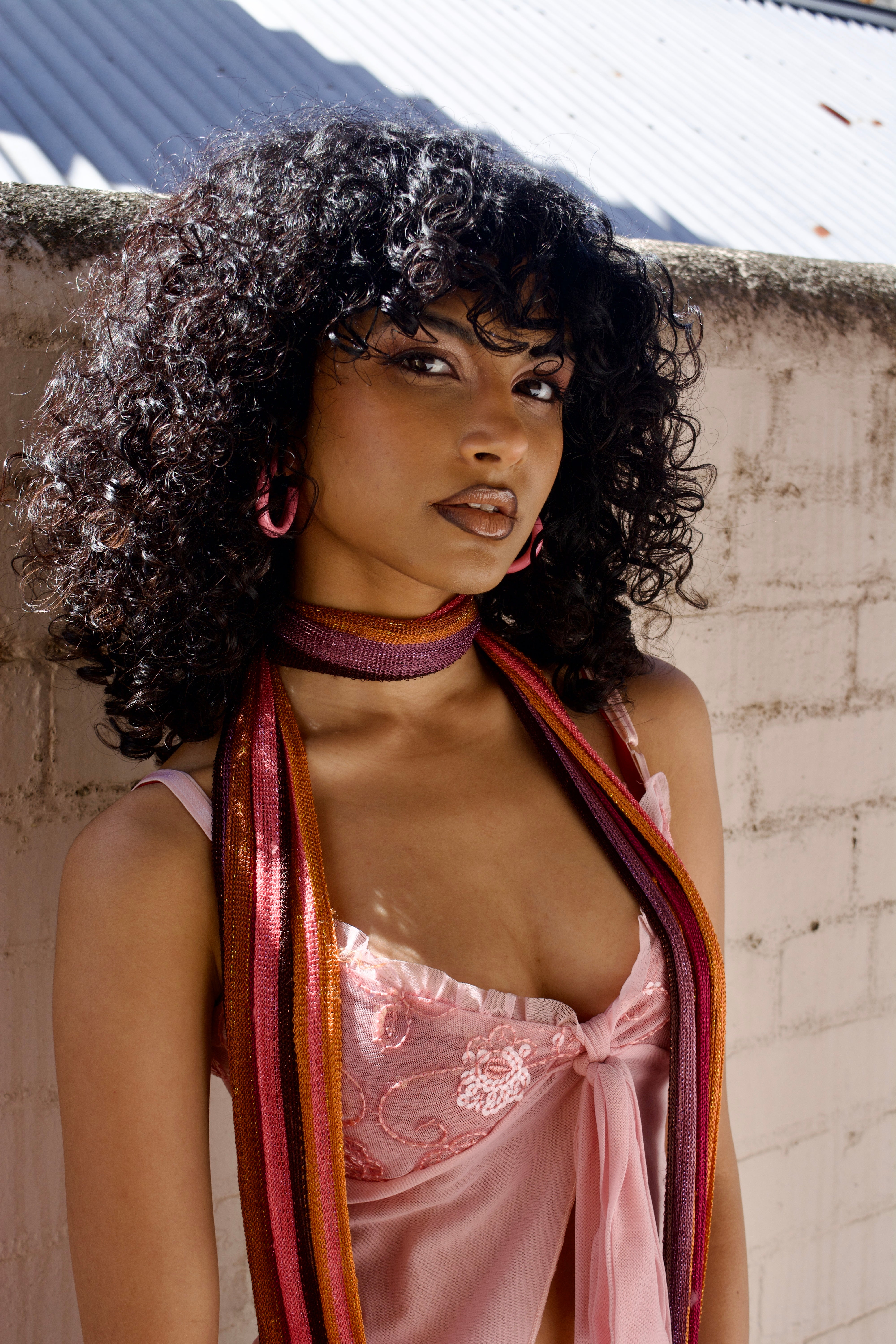 Woman wearing a pink lace top and colorful scarf against a white wall.