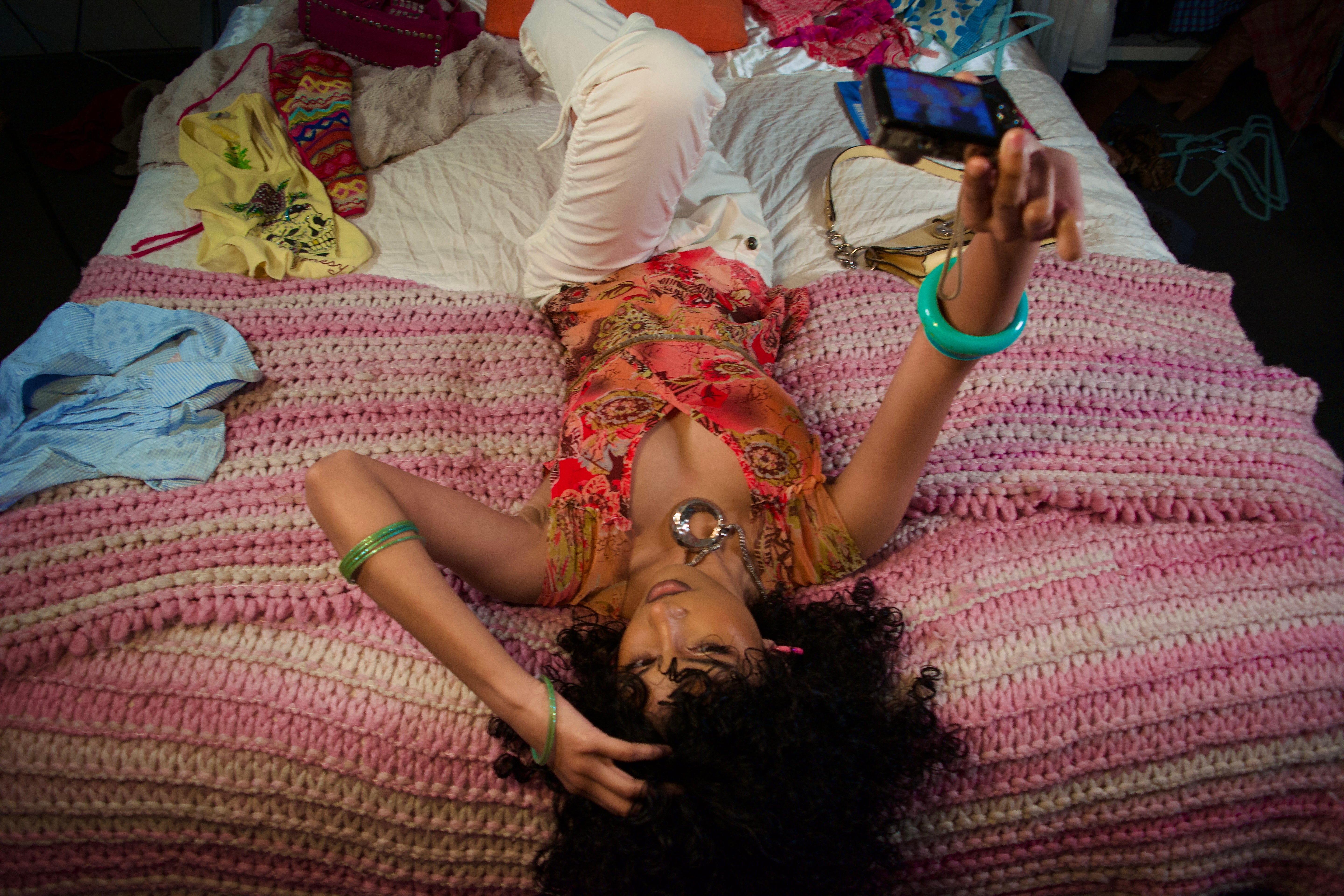 Person lying on a bed taking a selfie with a phone, wearing colorful clothing.