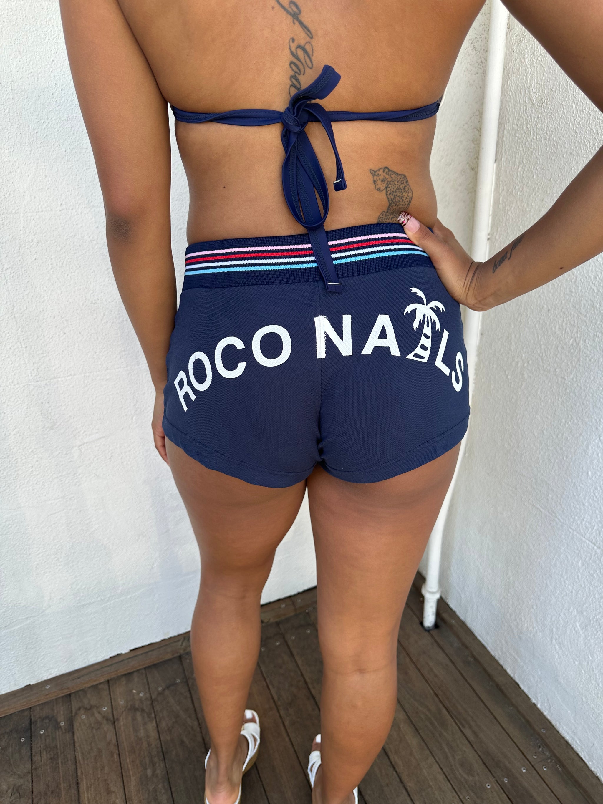 Woman Dressed in Y2K 00s Navy printed booty shorts featuring a navy base with “Roco Nails” printed across the bum. 