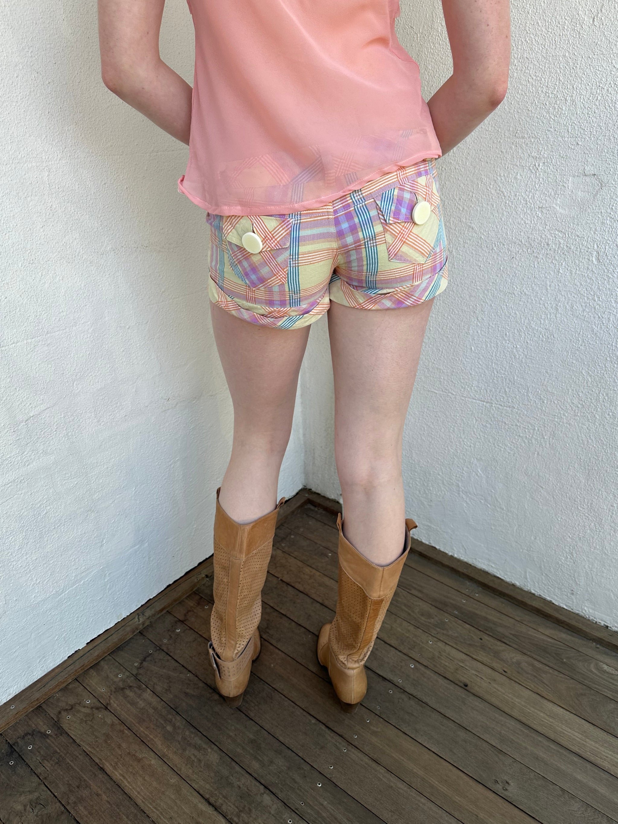 Woman Dressed in Y2K Pastel multicoloured tartan shorts featuring a fitted booty-style silhouette with a chunky pastel yellow button feature.