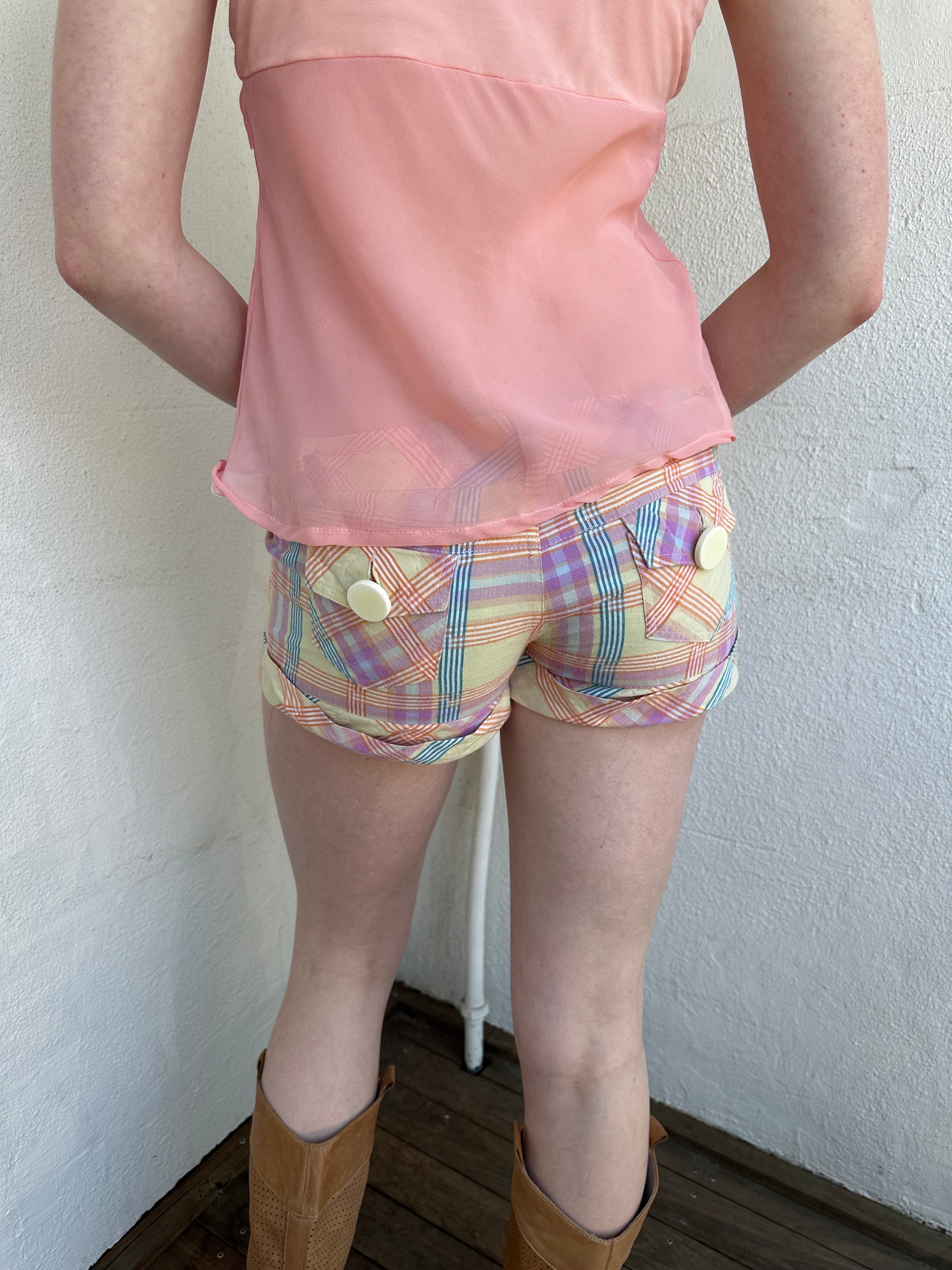 Woman Dressed in Y2K Pastel multicoloured tartan shorts featuring a fitted booty-style silhouette with a chunky pastel yellow button feature.
