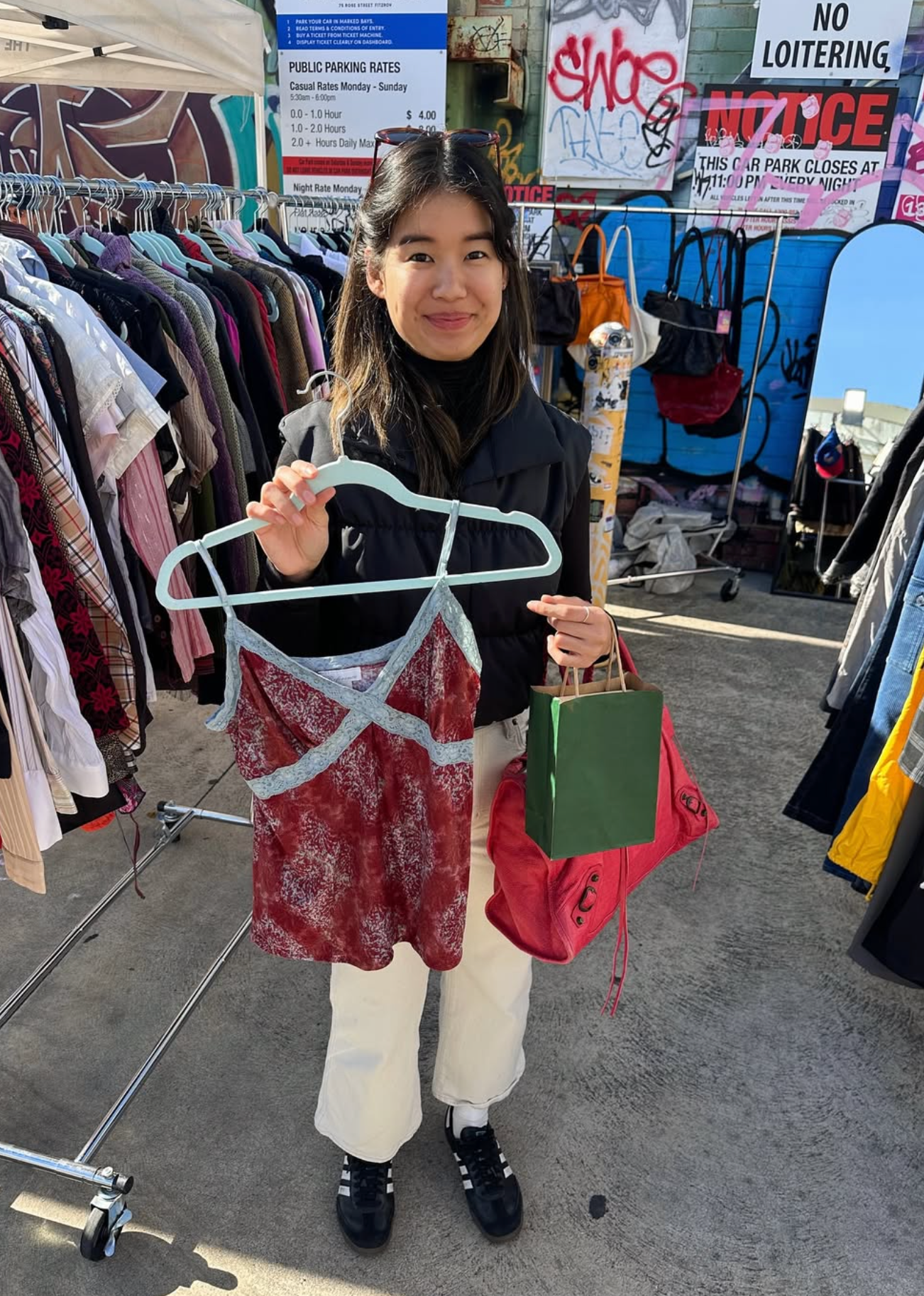 Person holding a hanger with a red top and a green bag at an outdoor market.