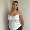 Woman Dressed in Y2K 00sWhite floral embroidered cami featuring a white and green embroidered flower design on the bust and a smooth satin body.