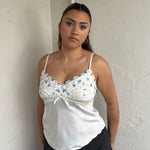 Woman Dressed in Y2K 00sWhite floral embroidered cami featuring a white and green embroidered flower design on the bust and a smooth satin body.