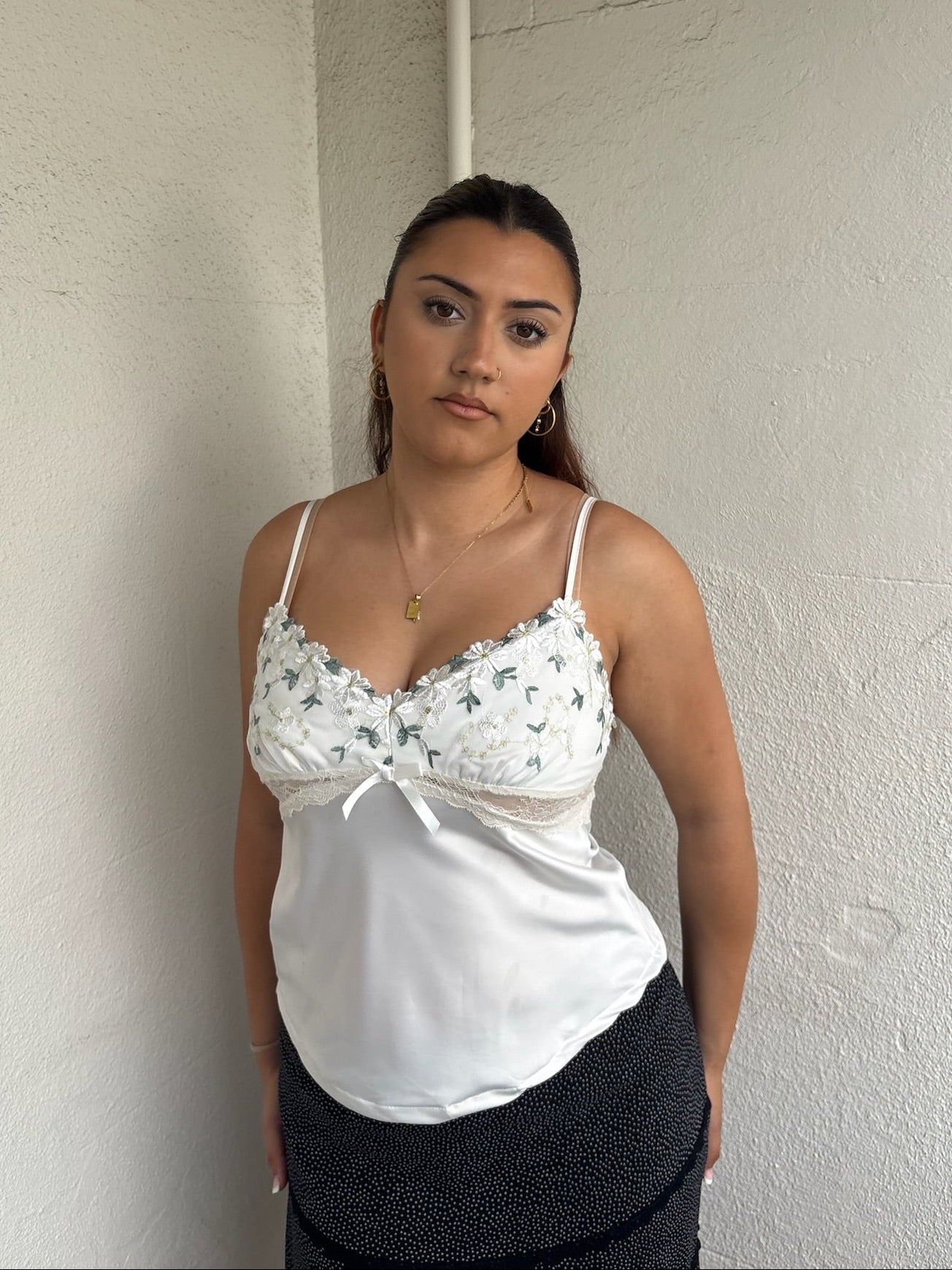 Woman Dressed in Y2K 00sWhite floral embroidered cami featuring a white and green embroidered flower design on the bust and a smooth satin body.