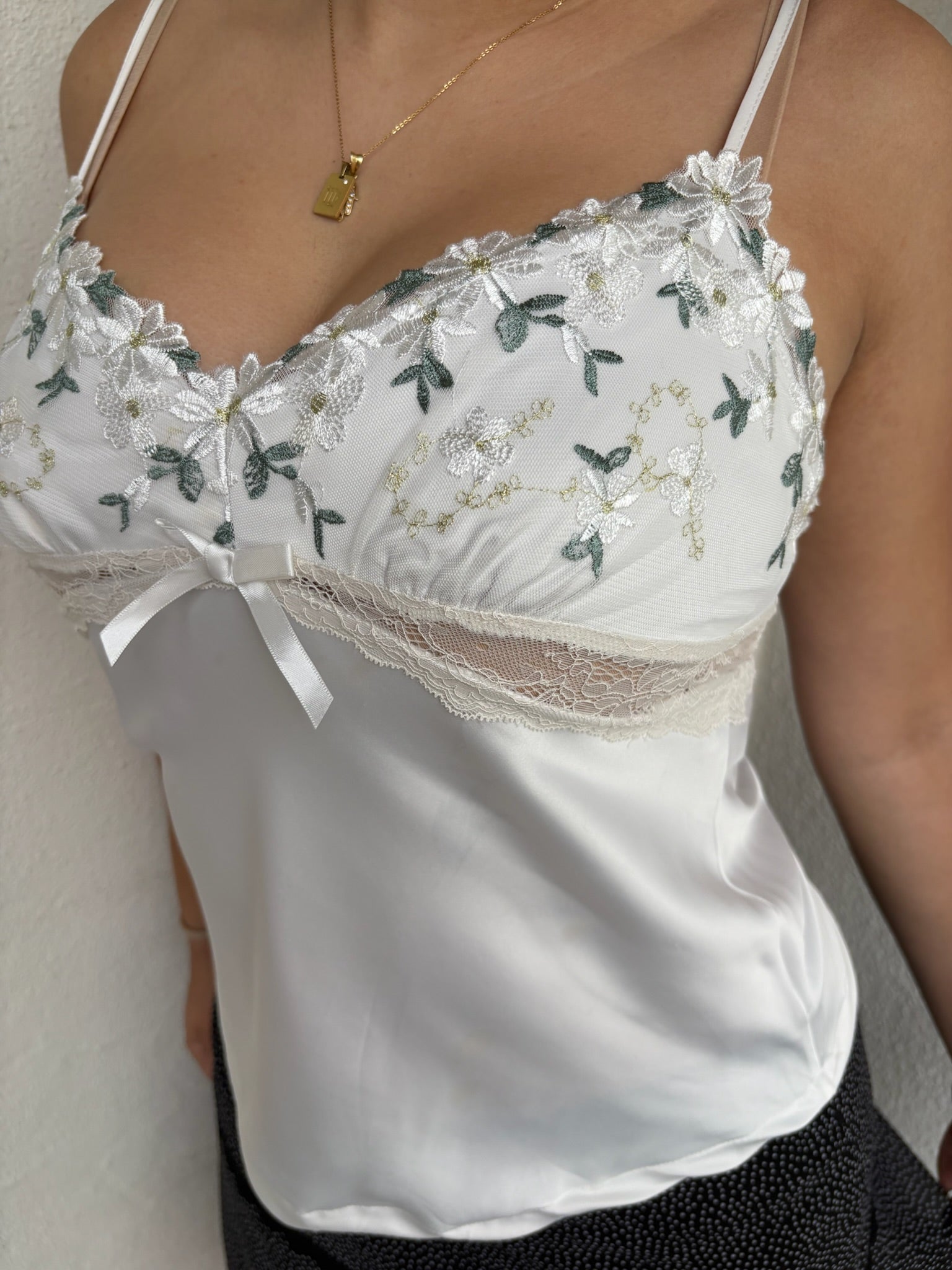 Woman Dressed in Y2K 00sWhite floral embroidered cami featuring a white and green embroidered flower design on the bust and a smooth satin body.