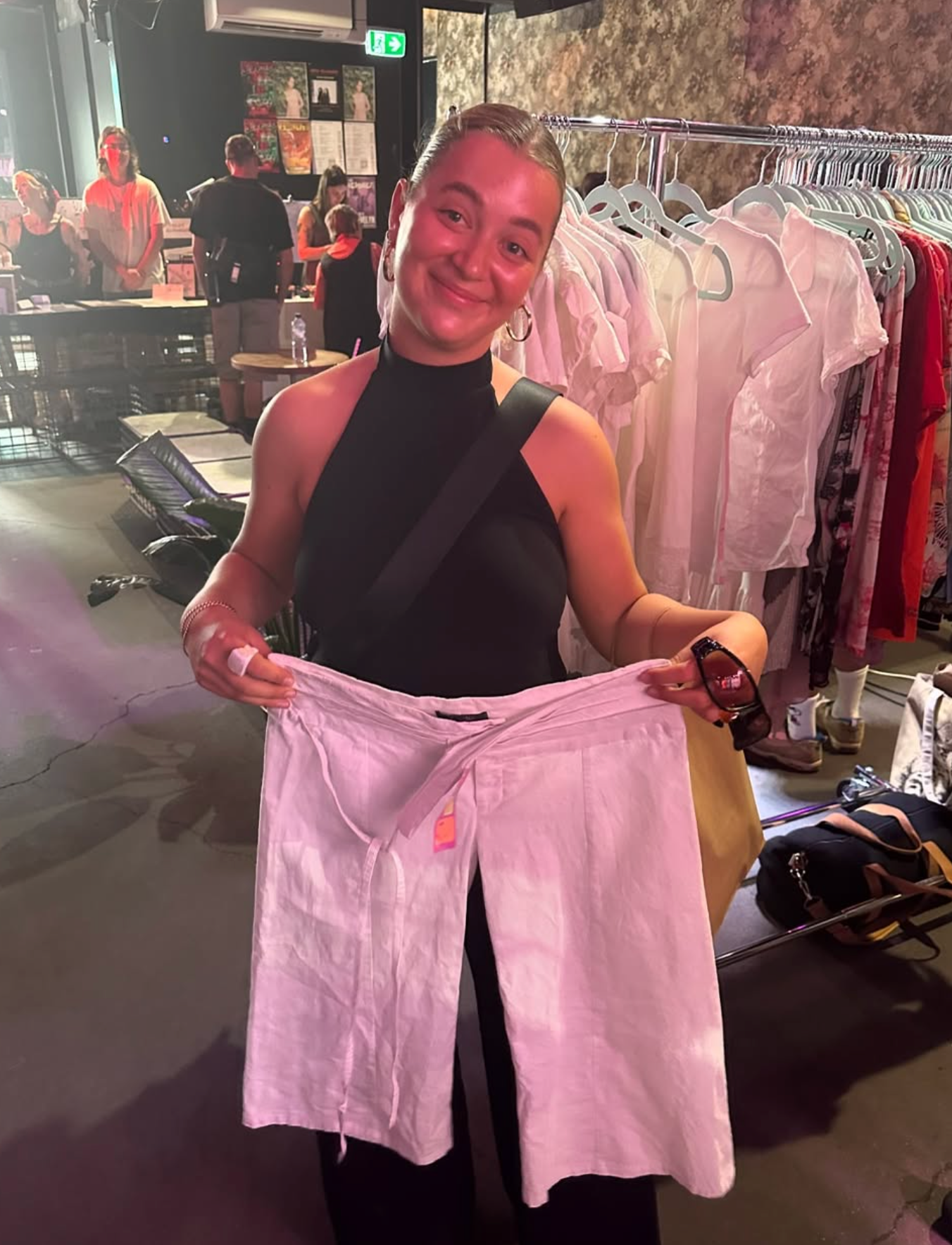 Person holding white pants in a market with racks of clothes in the background.