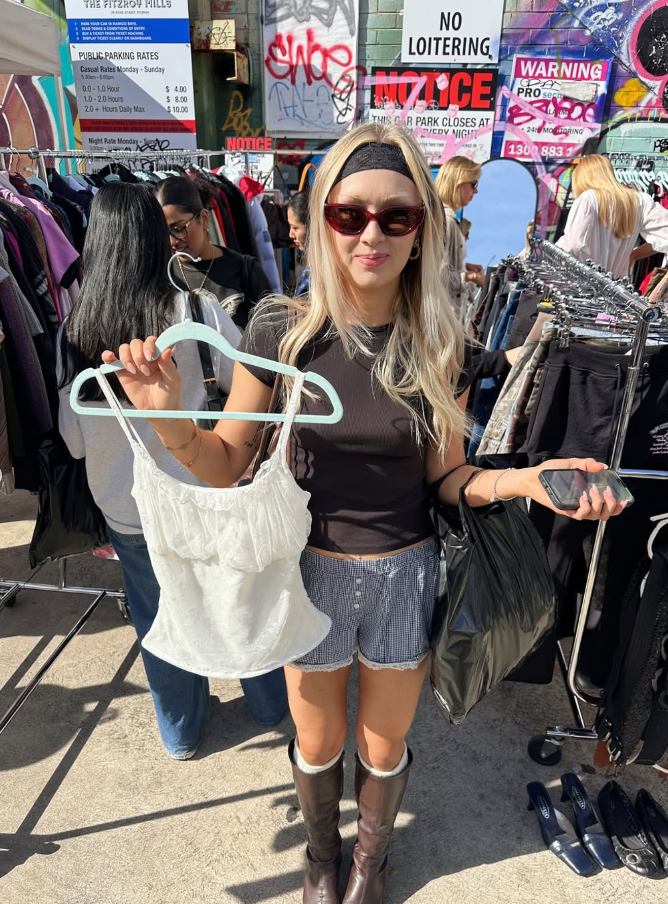 Woman holding a white top on a hanger in an outdoor market setting with various signs and clothing racks.