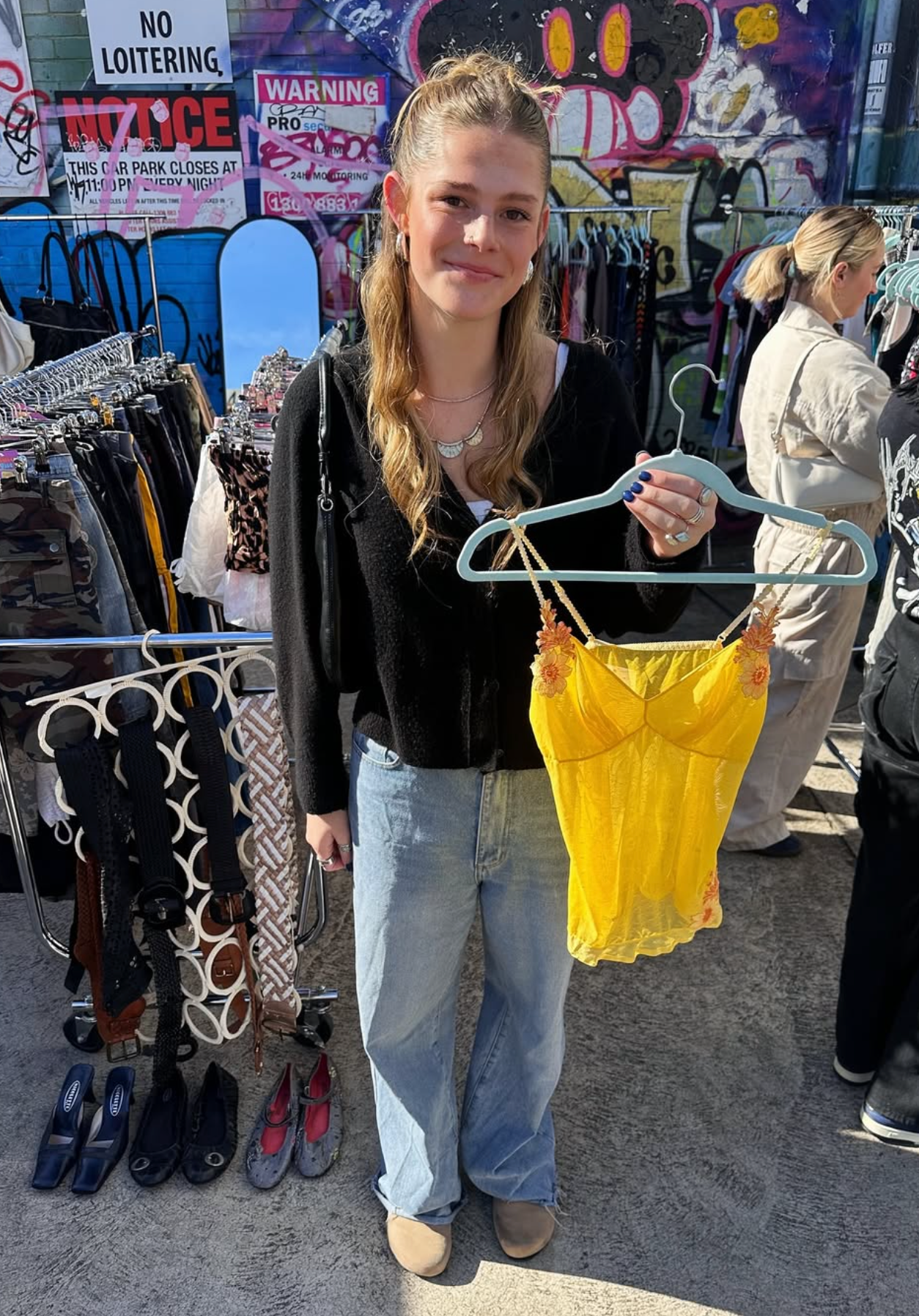 Person holding a yellow top on a hanger at a market with colourful signs in the background.